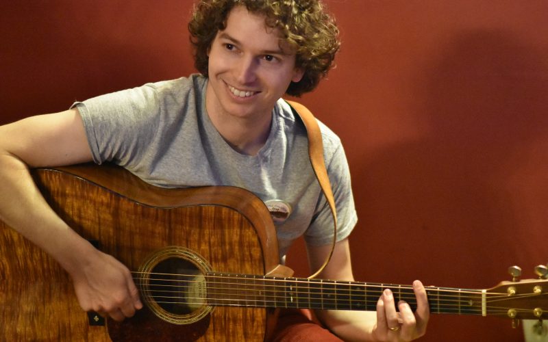 Owen Morrison, a man with curly brown hair is wearing a grey tee-shirt and smiling. He is sitting and playing a dark-colored guitar.