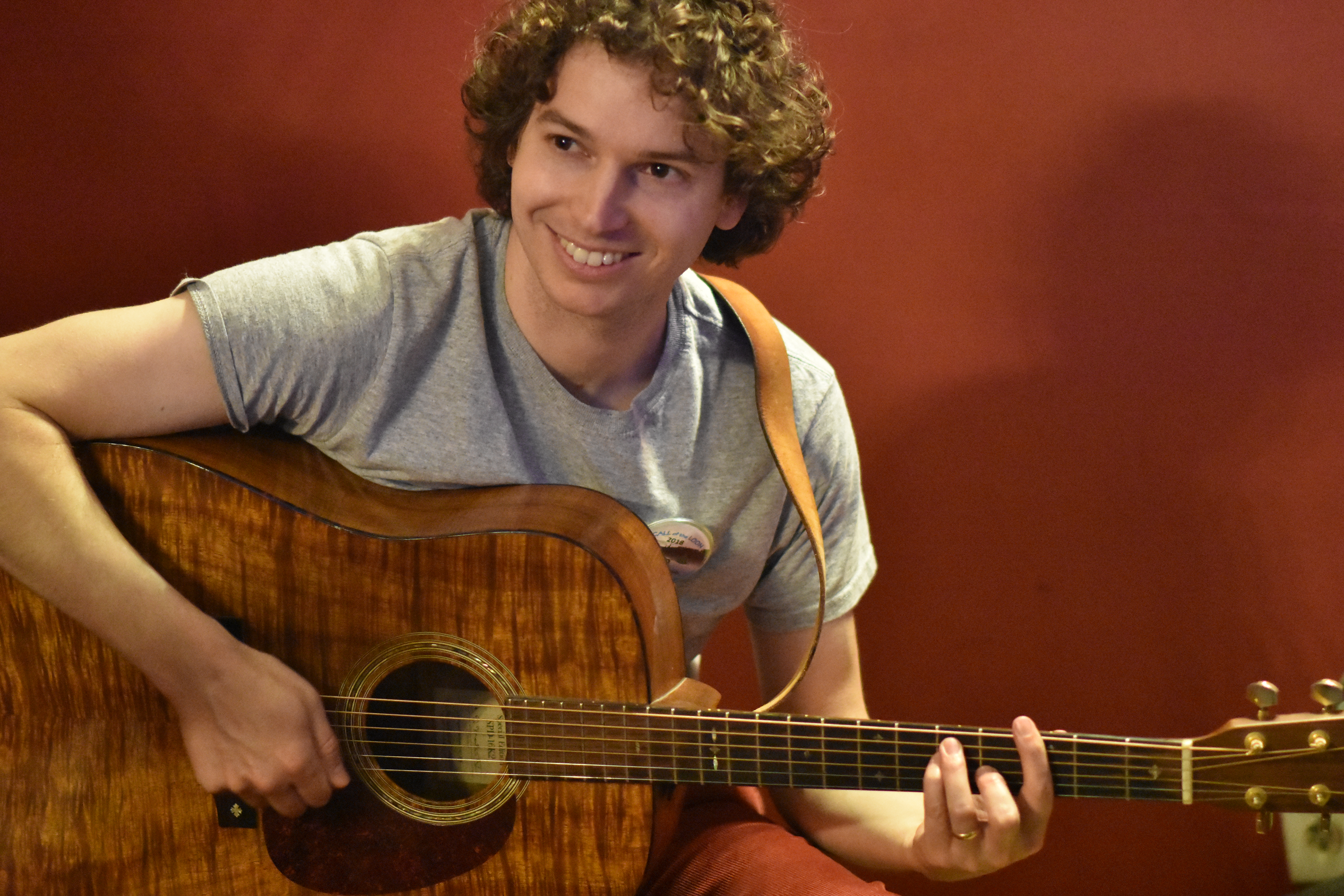 Owen Morrison, a man with curly brown hair is wearing a grey tee-shirt and smiling. He is sitting and playing a dark-colored guitar.